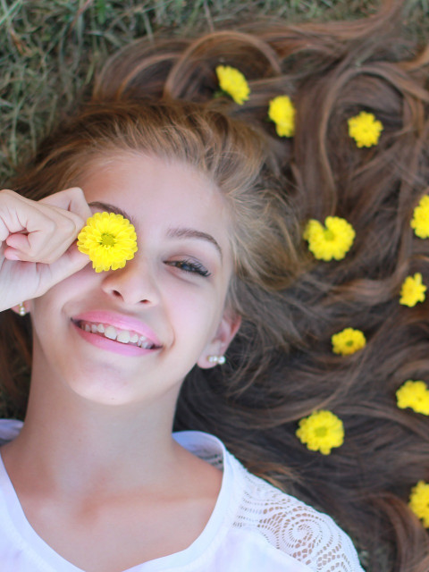 lächelnde Frau mit weißen T-Shirt und langen Haaren mit gelben Blüten im Gras