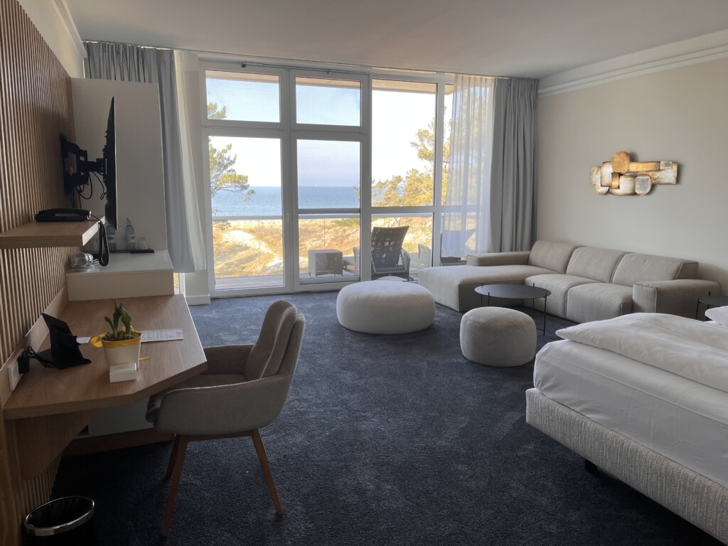 Suite im Strandhotel Fischland, Hauptraum mit Doppelbett, Couch, TV und Blick auf die Ostsee bei Sonnenschein
